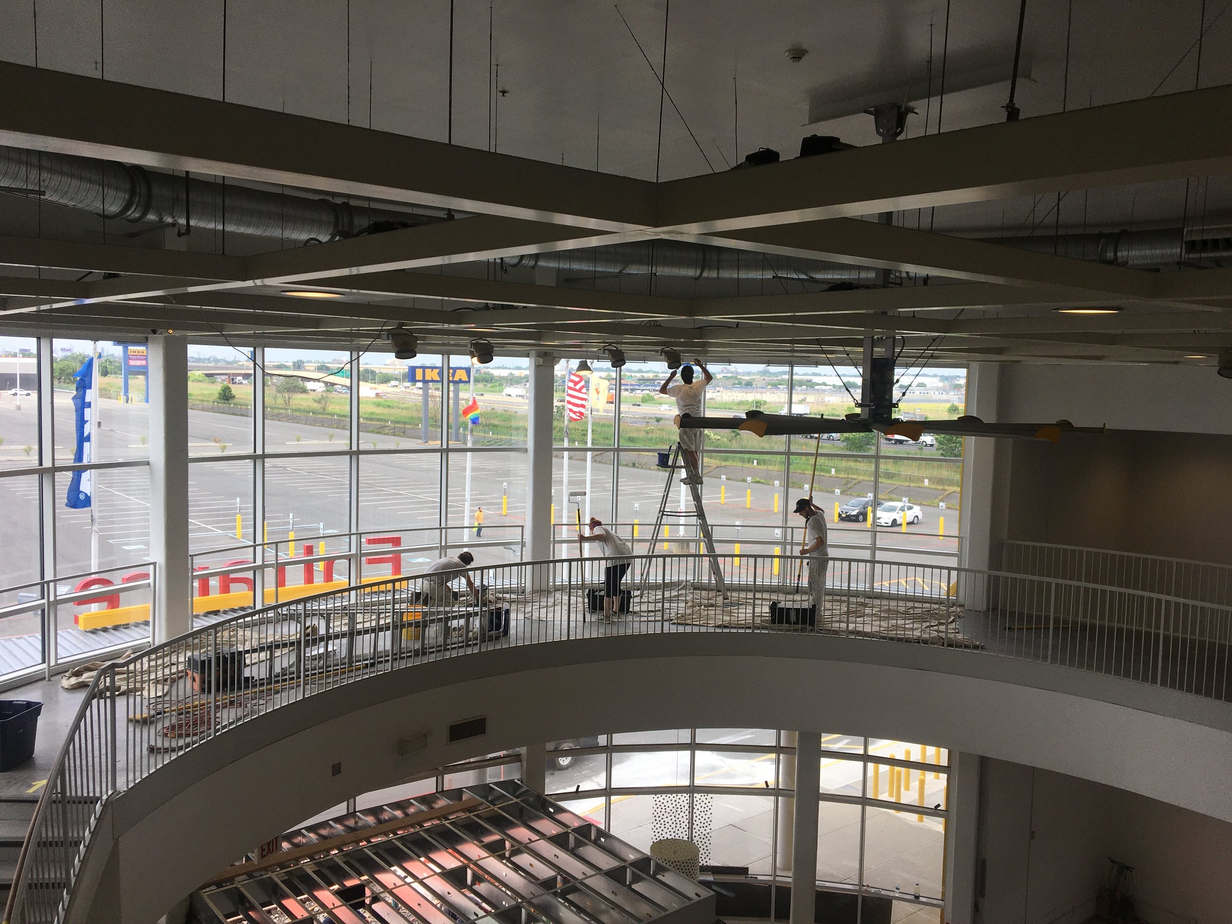 Workers on a ladder installing or cleaning lighting fixtures inside a large commercial building with a view of an empty parking lot outside through large windows.