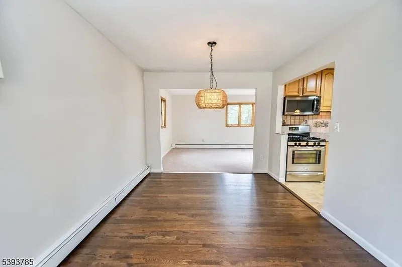 Empty living room and dining area with hardwood floors and a hanging wicker light fixture, opening to a kitchen with wooden cabinets and stainless steel appliances.