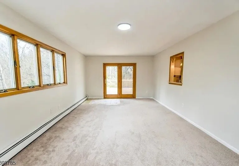 Empty living room with large windows, a sliding glass door leading outside, beige carpet, and white walls.