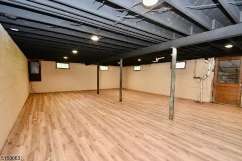 Empty basement with wood flooring, black ceiling with exposed beams, and small windows near the ceiling, including a wooden door on the right side.