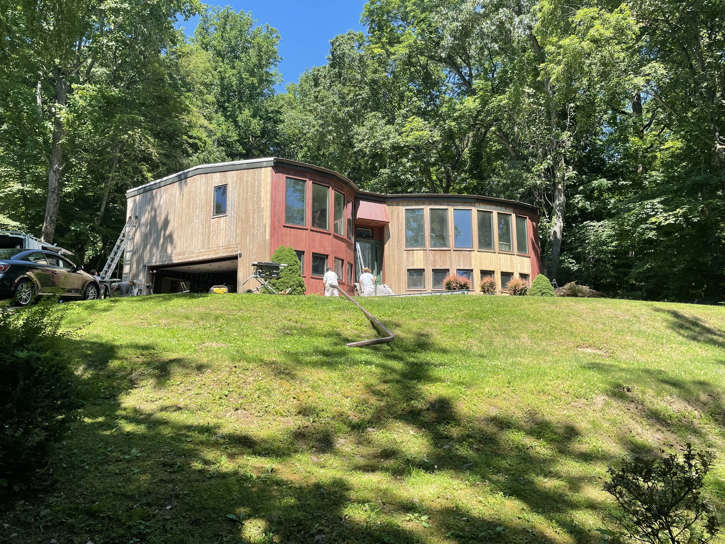 A house with rounded walls and large windows, set on a grassy hill surrounded by trees on a sunny day.