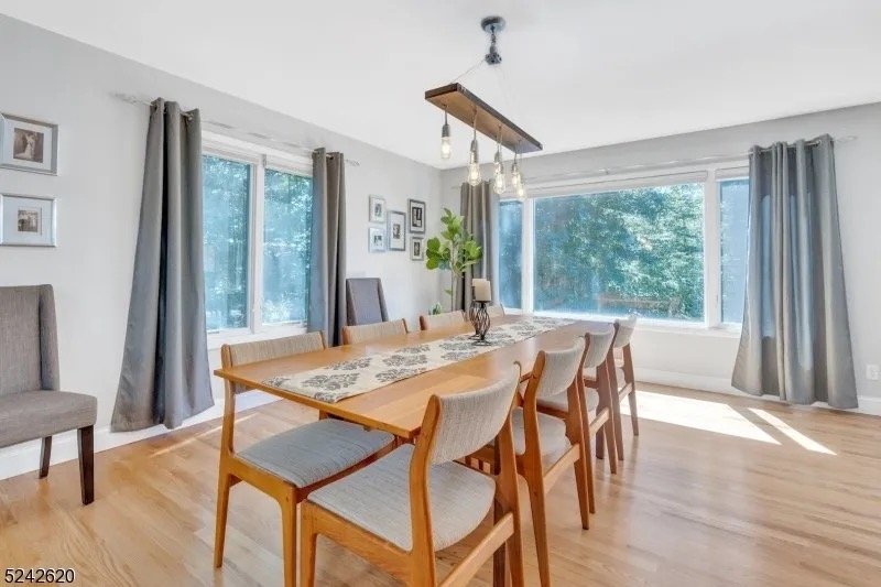 A bright dining room with a large wooden table surrounded by eight chairs, three with upholstered seats. The room has light wood flooring, white walls, large windows with gray curtains, and framed photos on the wall. A centerpiece with plants and a t