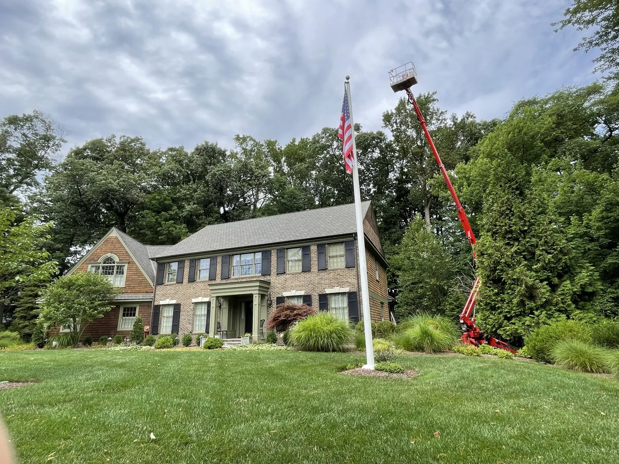 A two-story house with brick and wooden exterior, surrounded by a well-maintained lawn and shrubs. An American flag is on a pole in front. A hydraulic lift is extended toward the flagpole, likely for maintenance, with a cloudy sky overhead.