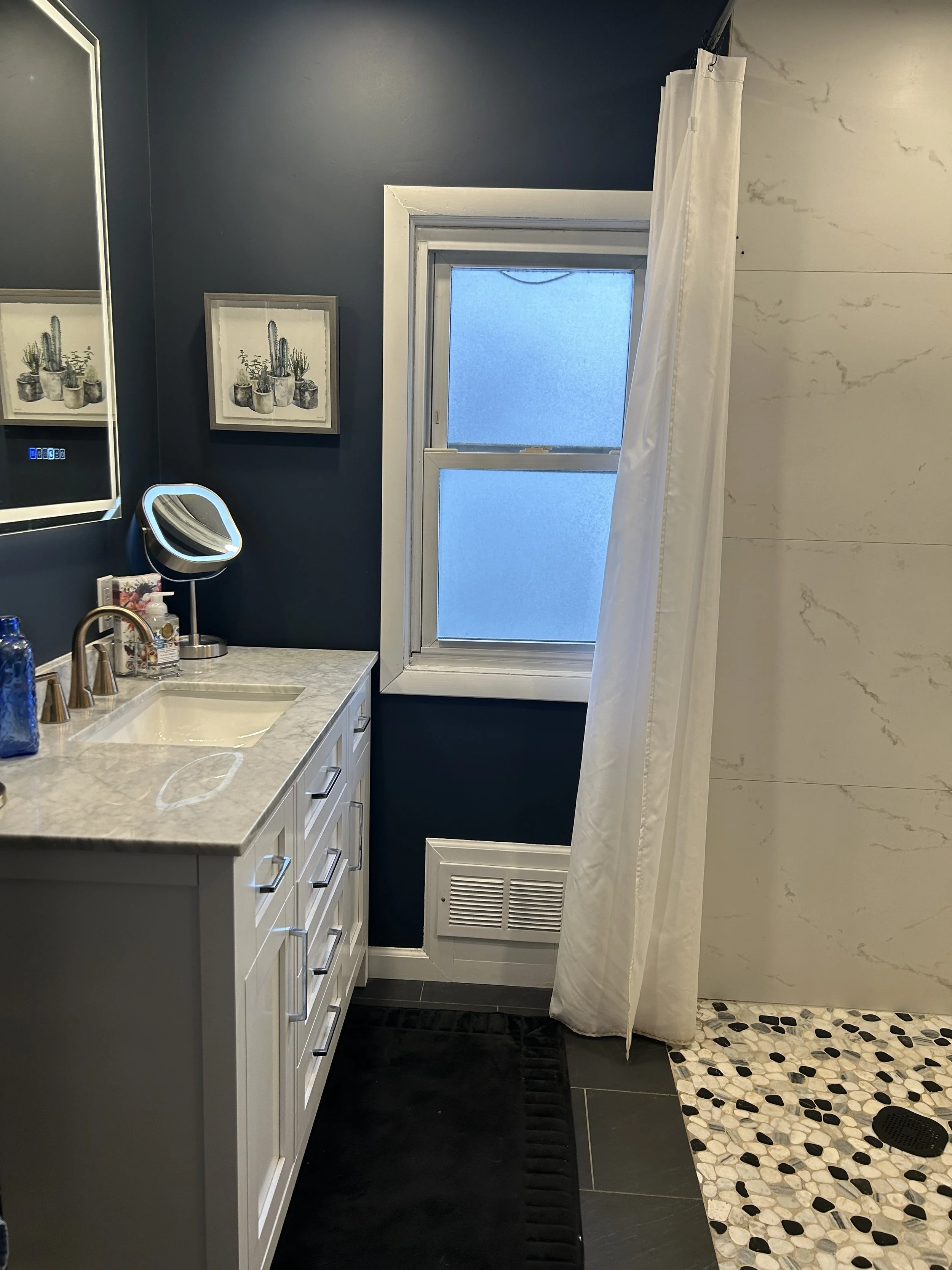 A bathroom with navy blue walls, a white marble countertop vanity with a sink, a small floating mirror, two framed cactus prints on the wall, a window with frosted glass, a shower with a white curtain, and a pebble-patterned shower floor.