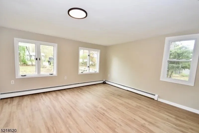 Empty living room with three windows, light-colored walls, light wood flooring, and baseboard heaters.