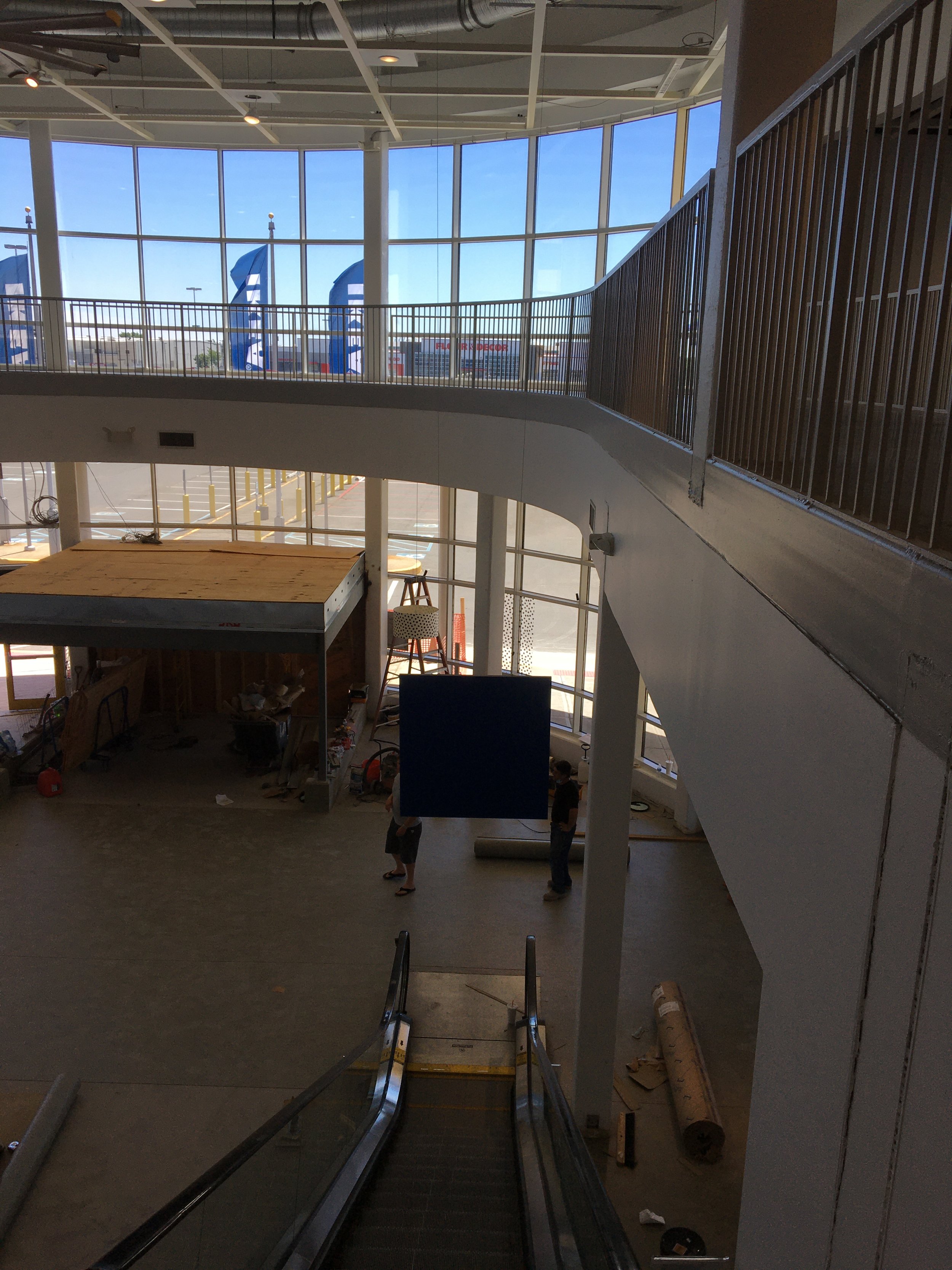 Interior view of a shopping mall or store under construction, with escalators, a large window, and construction materials and workers present.