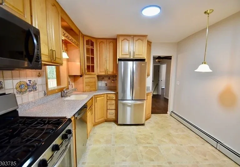 Kitchen with wooden cabinets, stainless steel refrigerator, black microwave, beige countertop, tile backsplash, and a window above the sink. A pendant light hangs from the ceiling, and a doorway shows a view into another room.