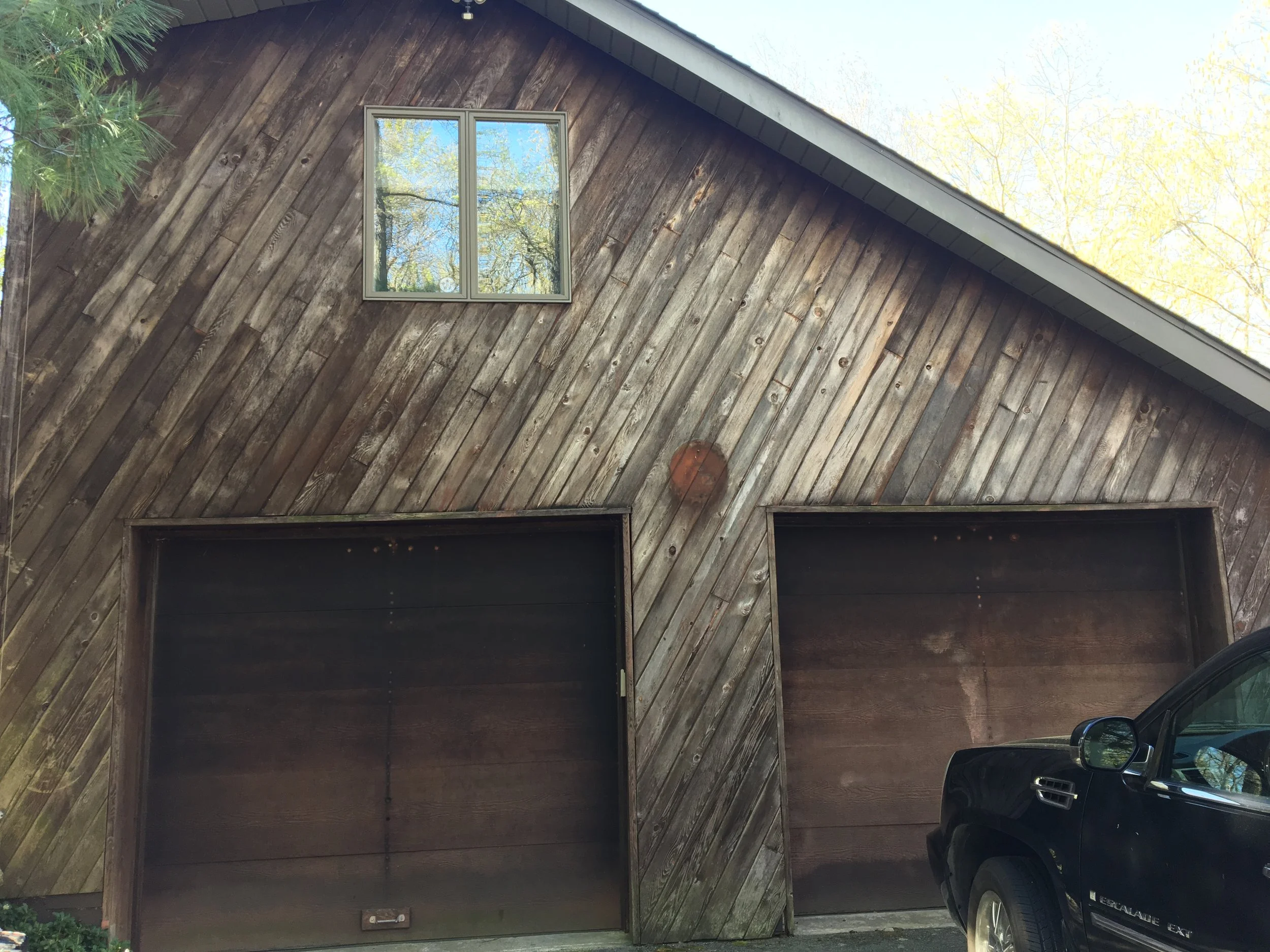 Front of a wooden garage with two large doors and a small window above, with a black vehicle parked in front on the right side.