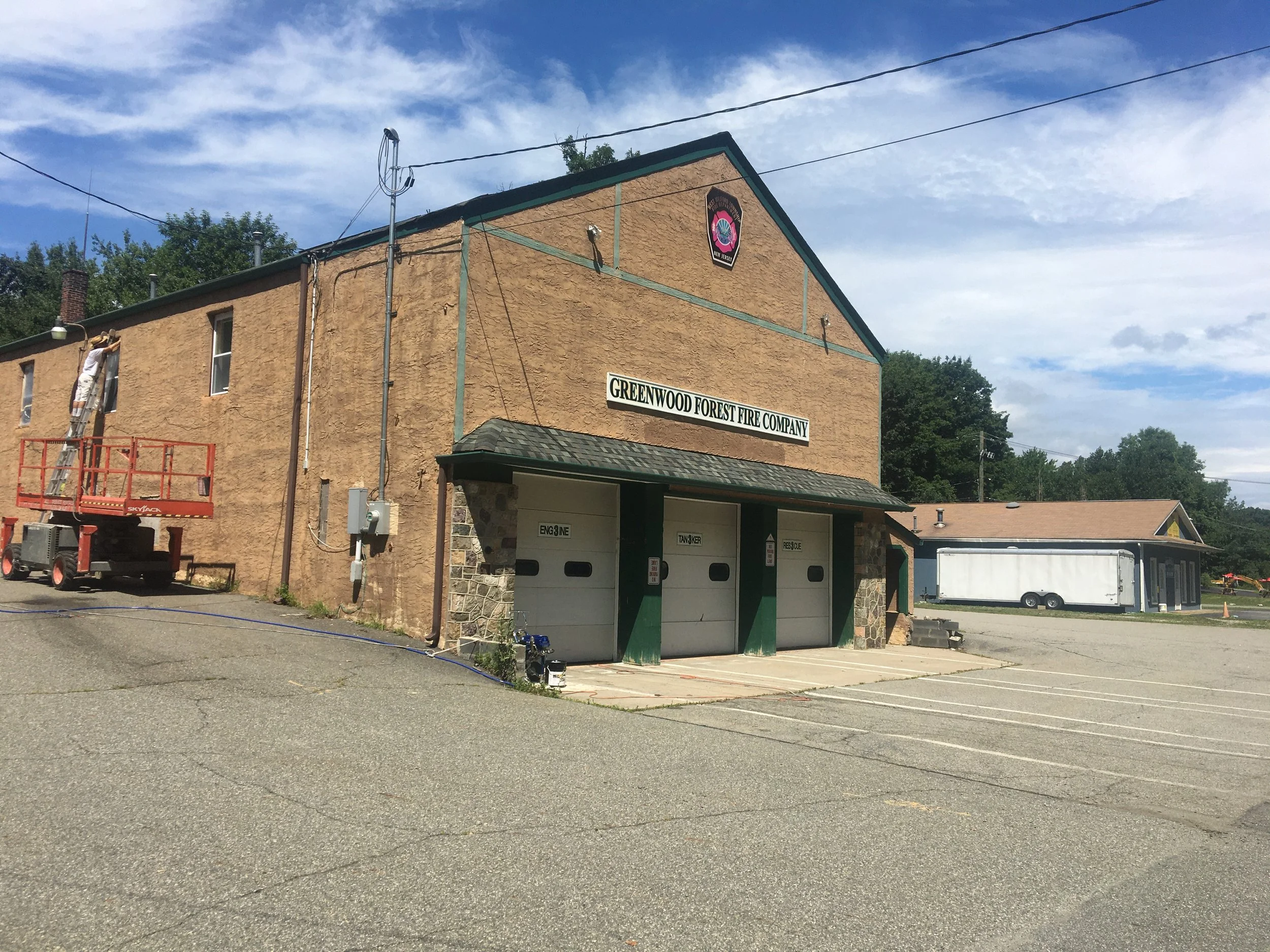 A brick building with a sign that reads 'Greenwood Forest Fire Company,' three garage doors, and a person on a lift working outside. The sky is partly cloudy.