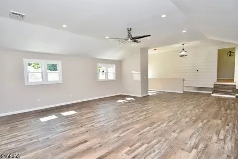 Empty living room with wood flooring, white walls, ceiling fan, windows, and stairs leading to an upper level.