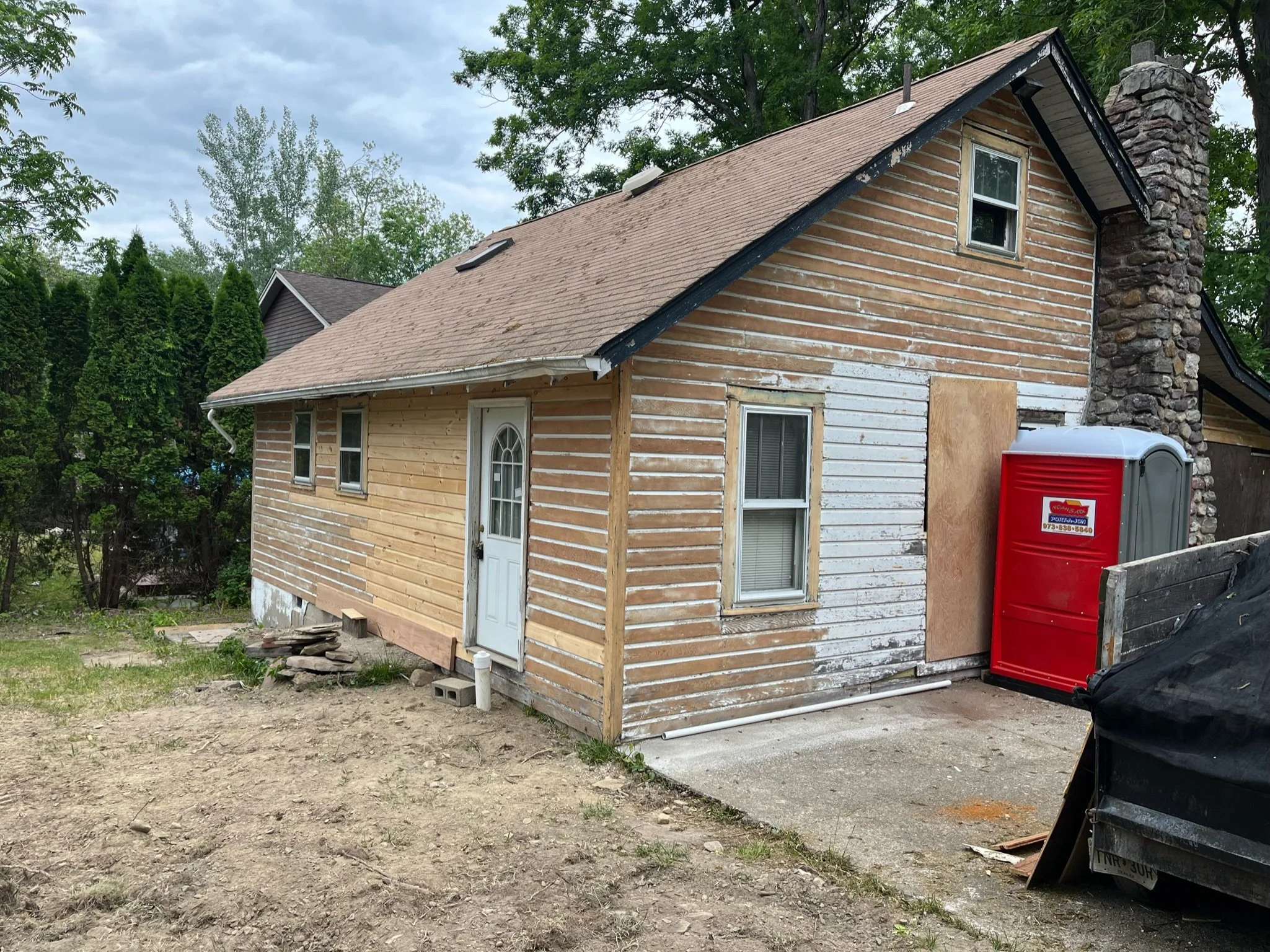A house under renovation with siding partially removed, showing different types of wood paneling. The house has a small upper window, a larger ground-floor window, and a front door. There is a red portable toilet next to the house, and the yard is mo