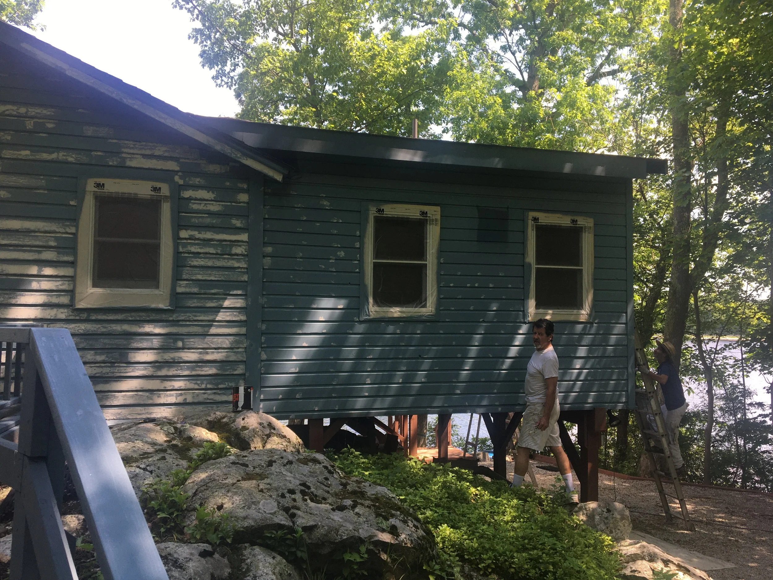Two people working on renovating a small house on stilts near a wooded area by a lake, with ladders and rocks in the foreground.