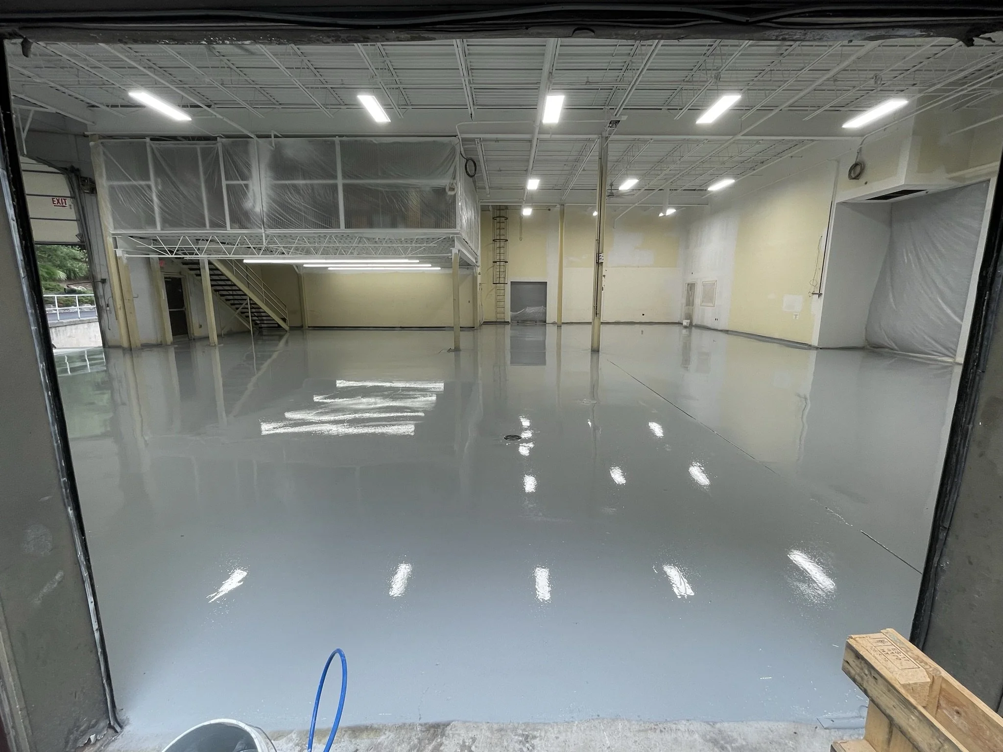 Empty warehouse with a new, shiny gray epoxy floor, high ceiling with bright fluorescent lights, and an upper level with metal stairs and railing.