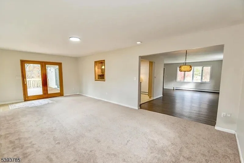 Empty living room with beige carpet, sliding glass door leading to a balcony, and an adjoining dining area with dark wood flooring and a hanging light fixture.