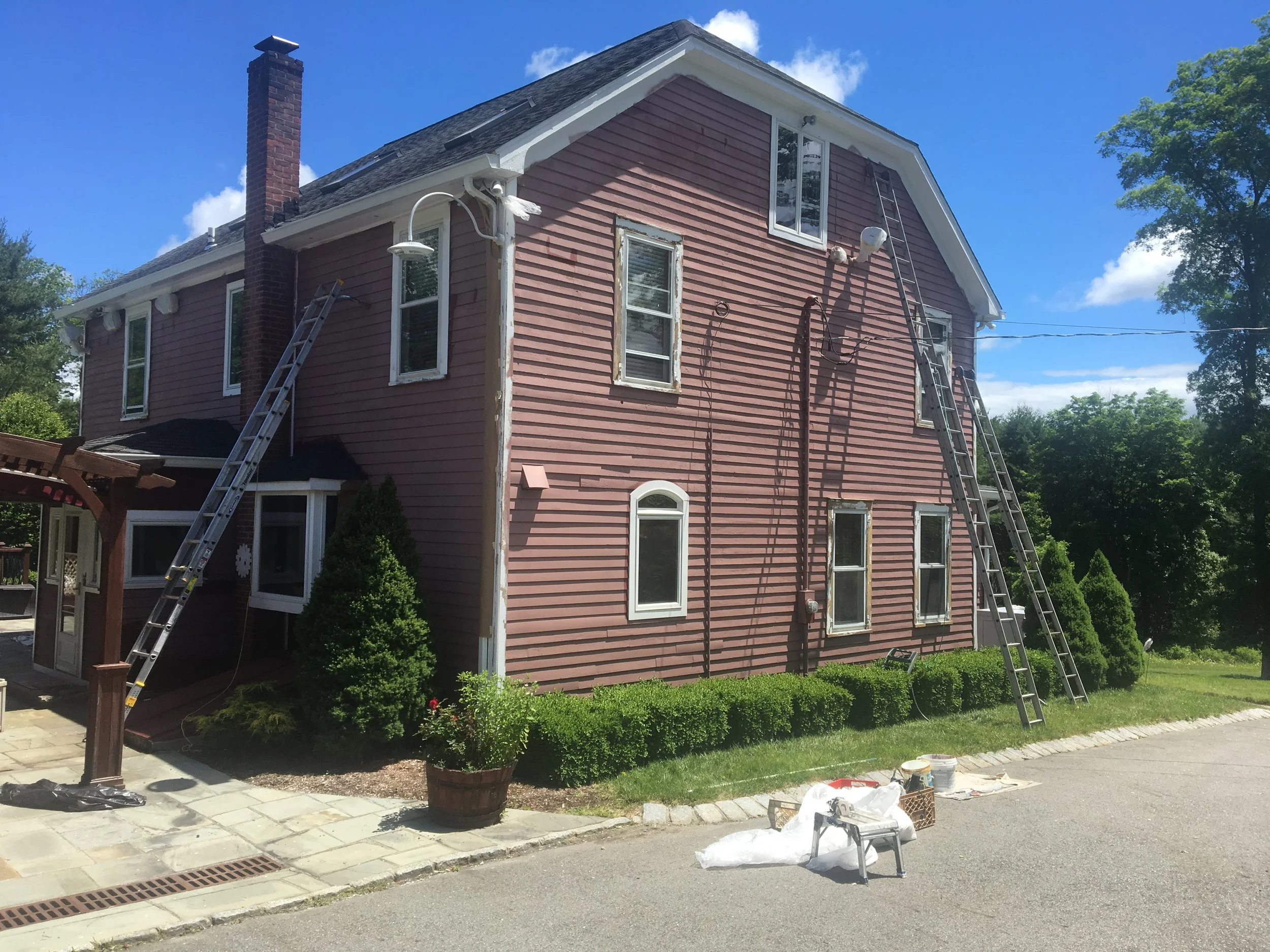 A two-story pink house with white trim is undergoing exterior painting or repair. Two ladders are leaning against the house, and there are two outdoor lights on the front and side walls. The house has several windows, a brick chimney, and is surround