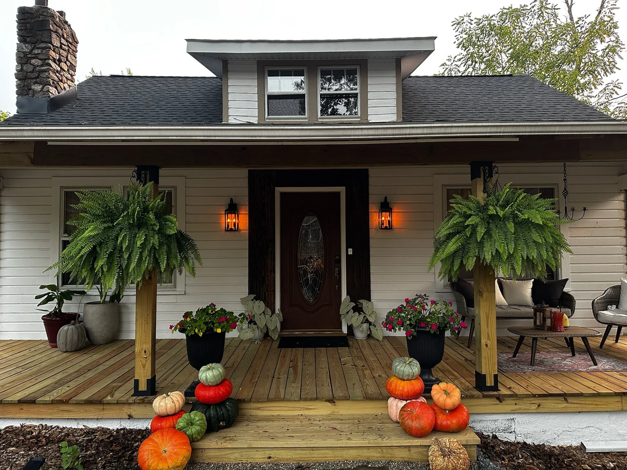 Front porch of a house decorated with pumpkins and potted plants, including two large hanging ferns, with a central wooden door and two warm outdoor wall lanterns.