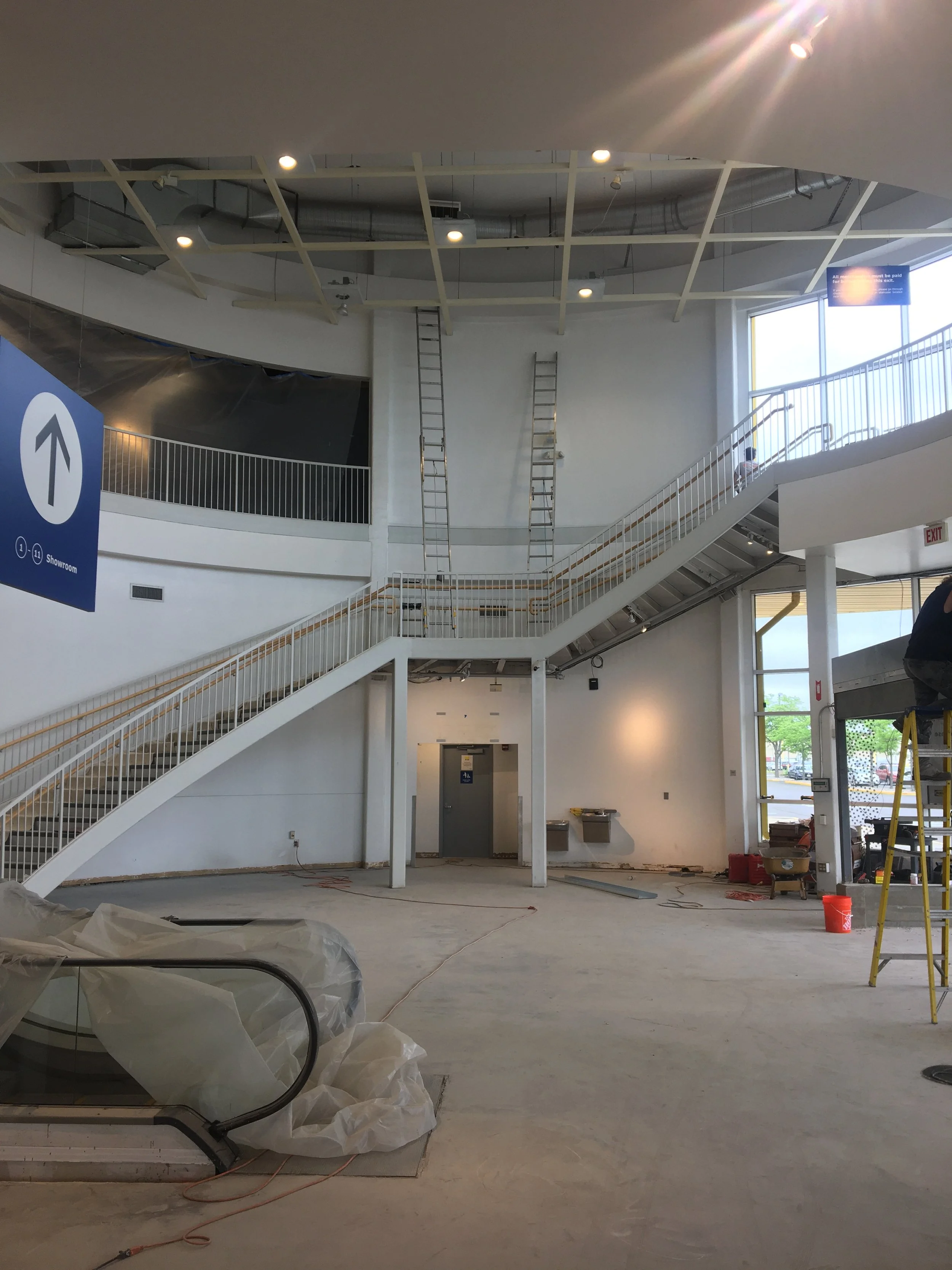 Interior of a building under construction, with a staircase, construction materials, and equipment. There is a blue sign with a white arrow pointing up, and a large window letting in natural light.