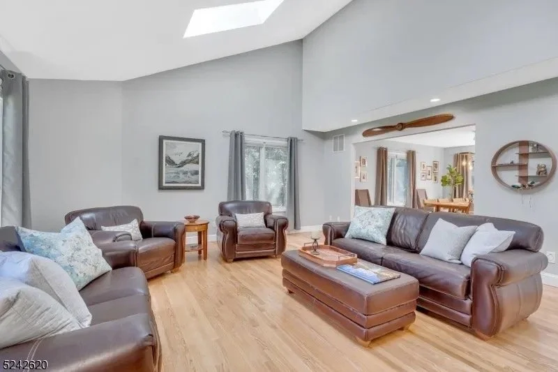 Spacious living room with light gray walls, hardwood floors, leather sofas, a wooden ottoman, and framed art, with windows and curtains allowing natural light.