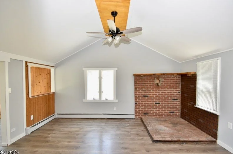 Empty living room with a brick fireplace, white walls, hardwood floor, ceiling fan, and windows with blinds.
