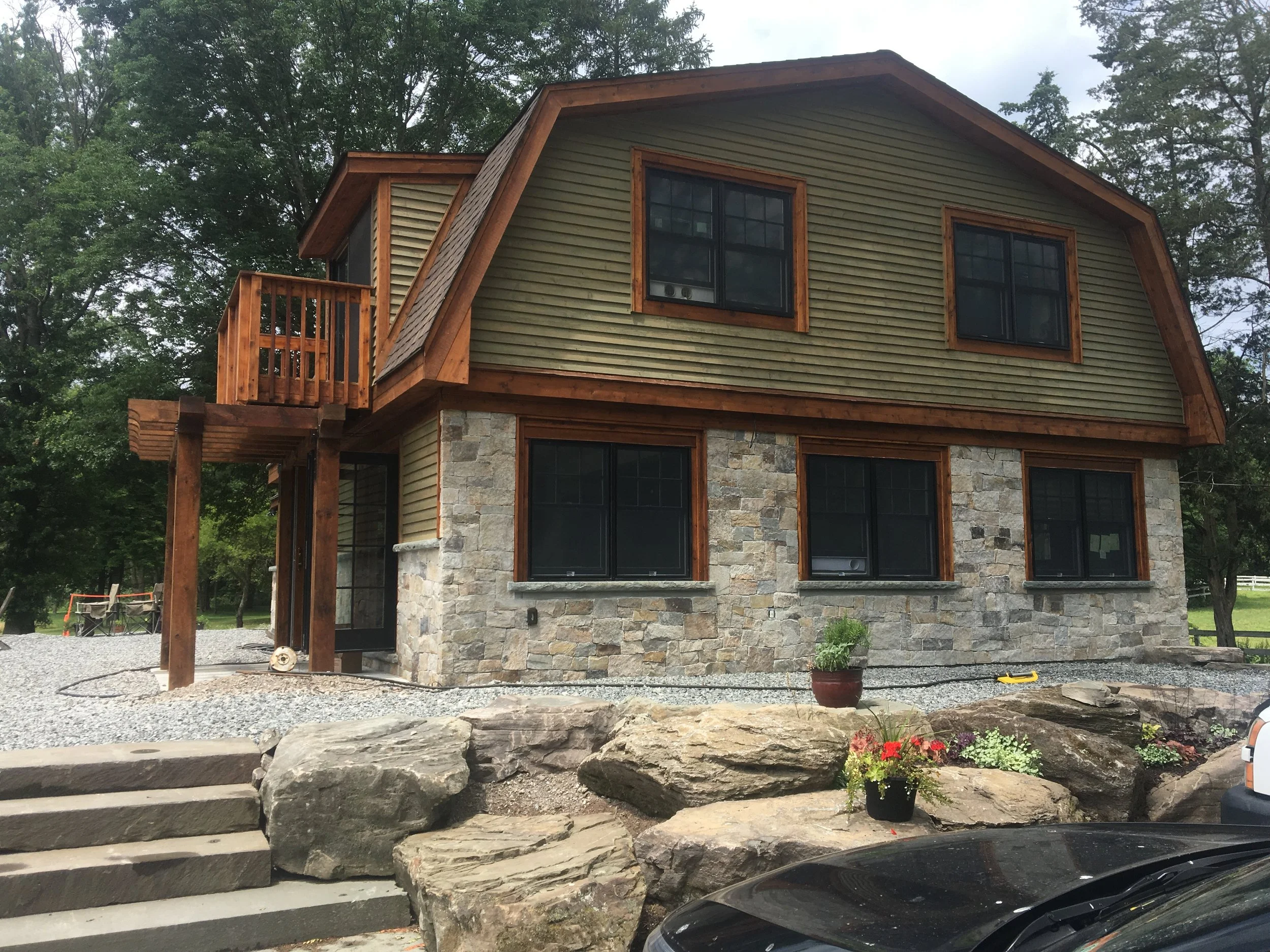 Front view of a two-story house with stone and wood exterior, multiple black-framed windows, a small balcony with wooden railing, stone steps leading up to the entrance, and landscaped rocks and flowers in front.