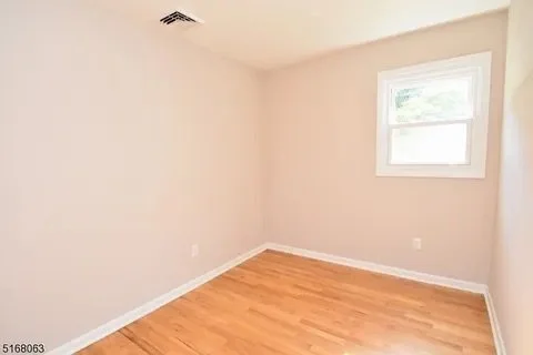 Empty room with beige walls, hardwood floor, white window, and ceiling vent.