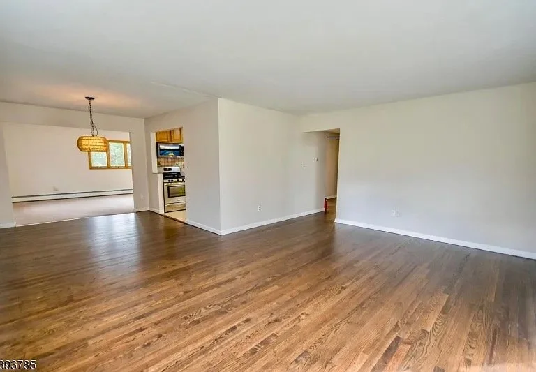 Empty living room with hardwood floors, white walls, and a view into an adjacent room with a hanging light fixture and a window.