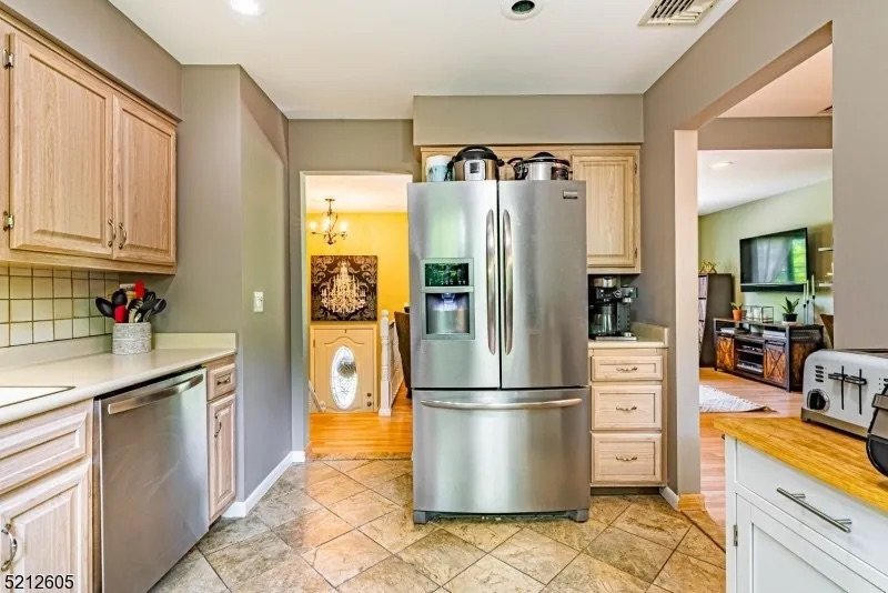 View of a modern kitchen with a stainless steel refrigerator, wooden cabinets, and beige tile flooring. Adjacent to the kitchen is a living room with a TV and a wooden console table. The background includes a laundry area with an open doorway.