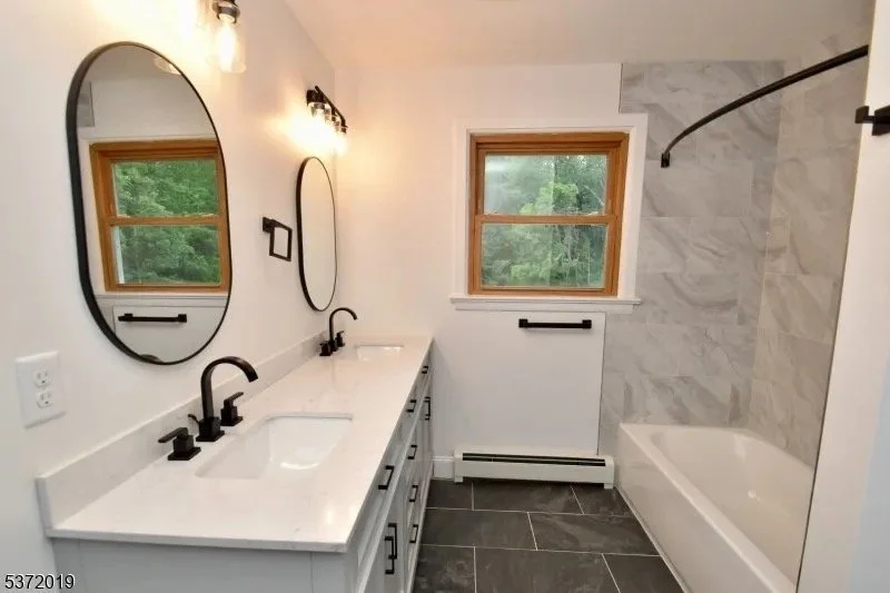 A bathroom with a double vanity with white countertop, two black faucets, and round black-framed mirrors. There are two windows with wooden trim, a black towel bar, and a bathtub with a marble tile surround. The floor is covered with dark tiles.