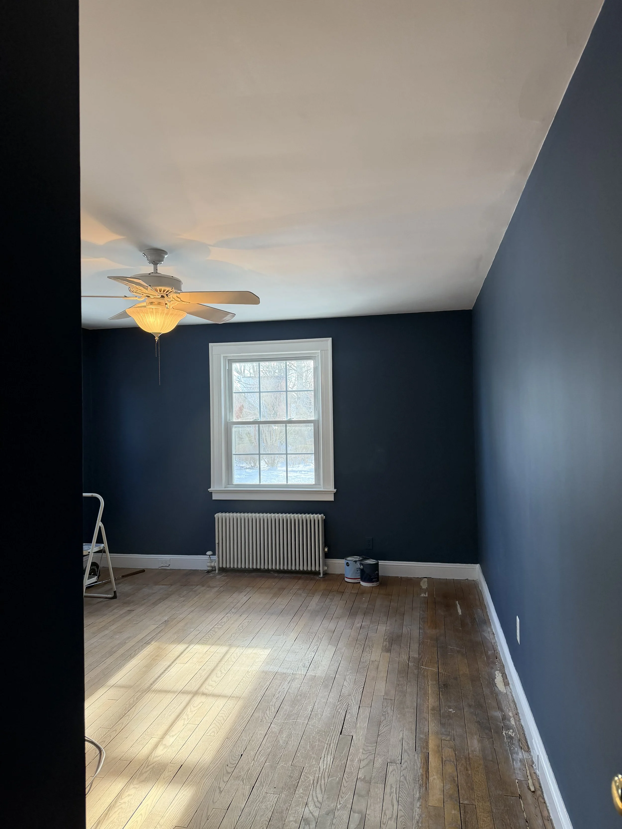 Empty room with navy blue walls, wooden floor, window, radiator, ceiling fan with light, and paint cans.