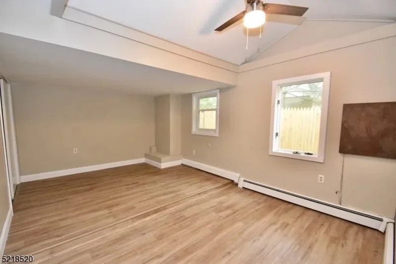 Empty room with hardwood floors, beige walls, two windows facing outside, ceiling fan with light, baseboard heater, and a small platform in the corner.