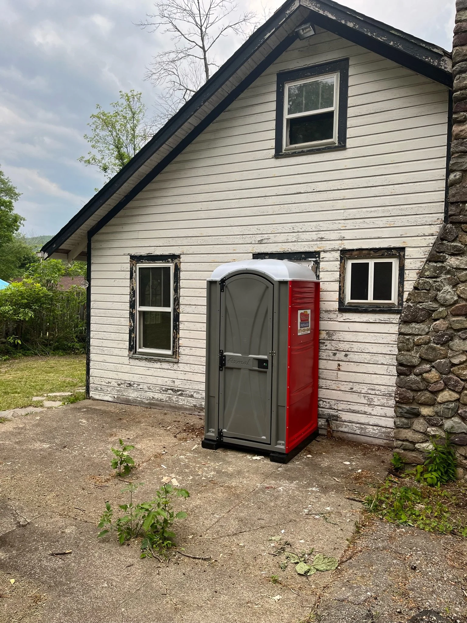A portable toilet situated on a concrete driveway outside a white house with black trim and a stone chimney, surrounded by some greenery and trees under a cloudy sky.