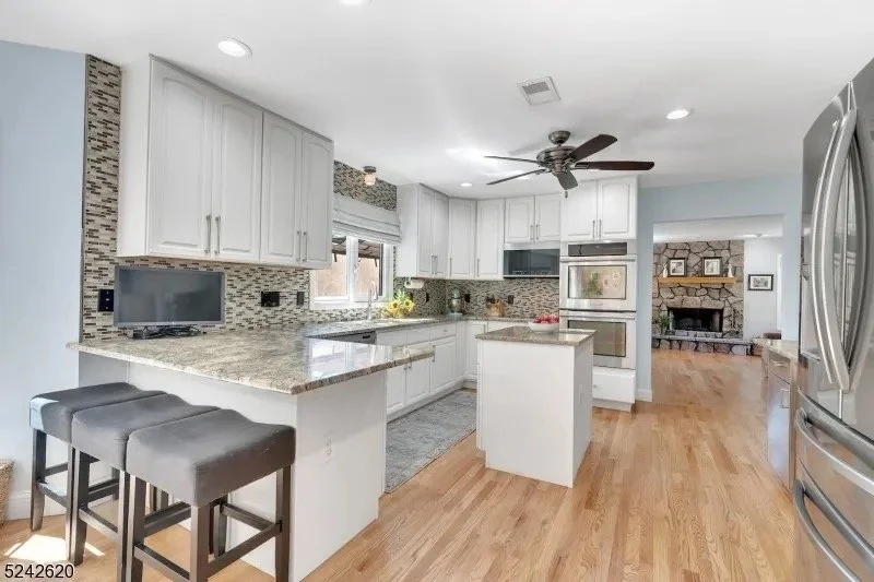 Modern kitchen with white cabinets, granite countertops, stainless steel appliances, and a small island with bar stools. The room has a hardwood floor and a view into a living area with a stone fireplace.