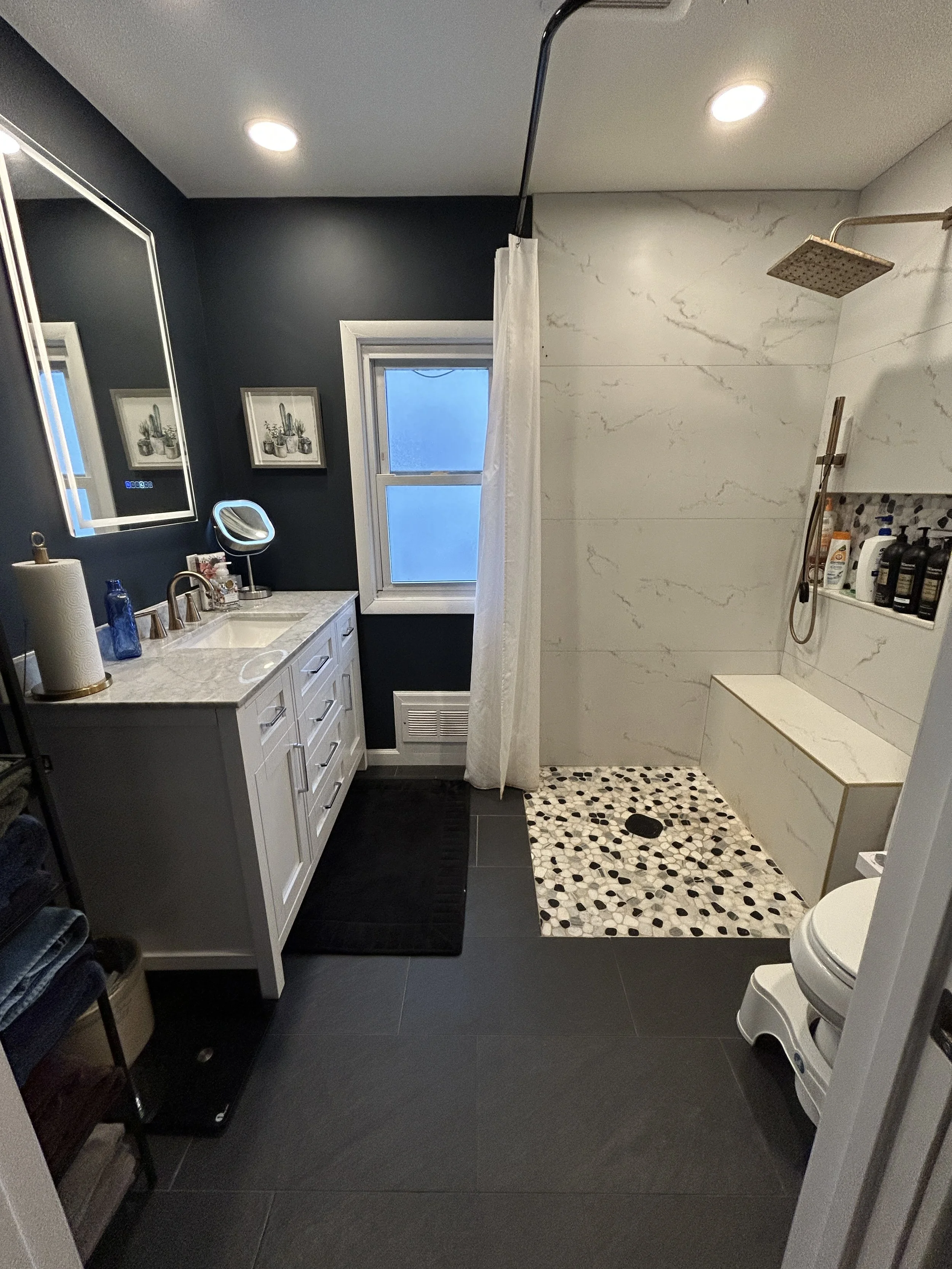 Bathroom with dark tile floor, white vanity with marble countertop, mirror, two framed cactus pictures, window, shower area with white marble walls, pebble tile floor, and built-in bench, showerhead, and toiletries.