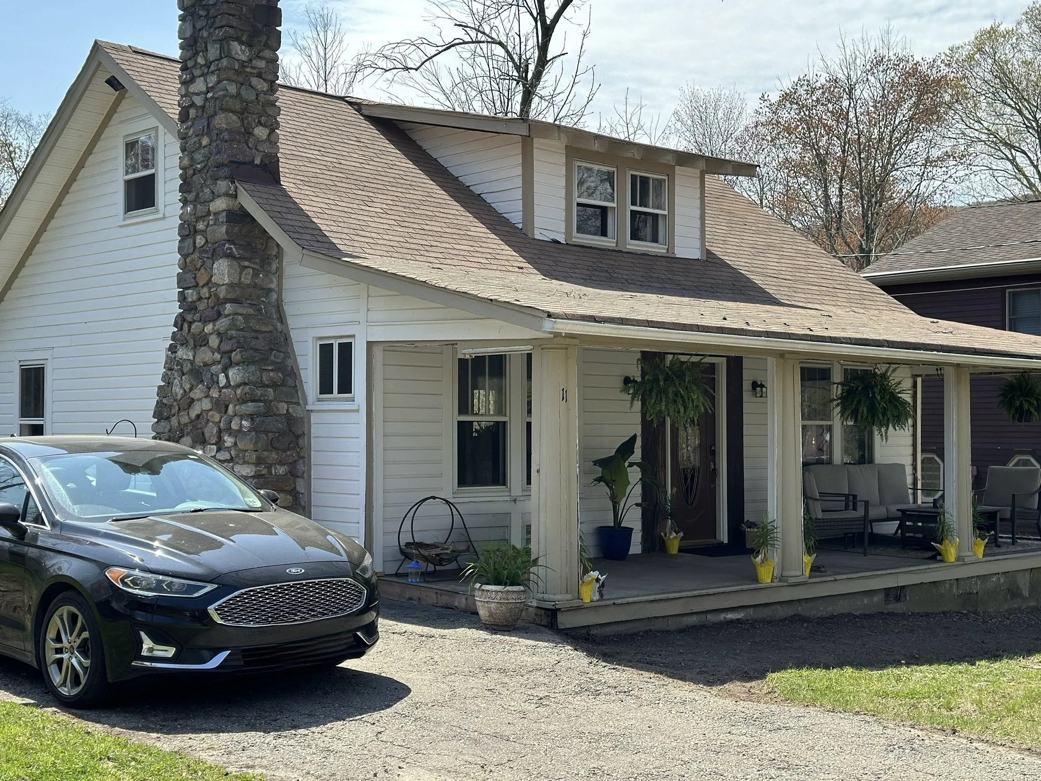 A house with a stone chimney, white siding, and a small porch with outdoor furniture and plants, parked gray car in front.