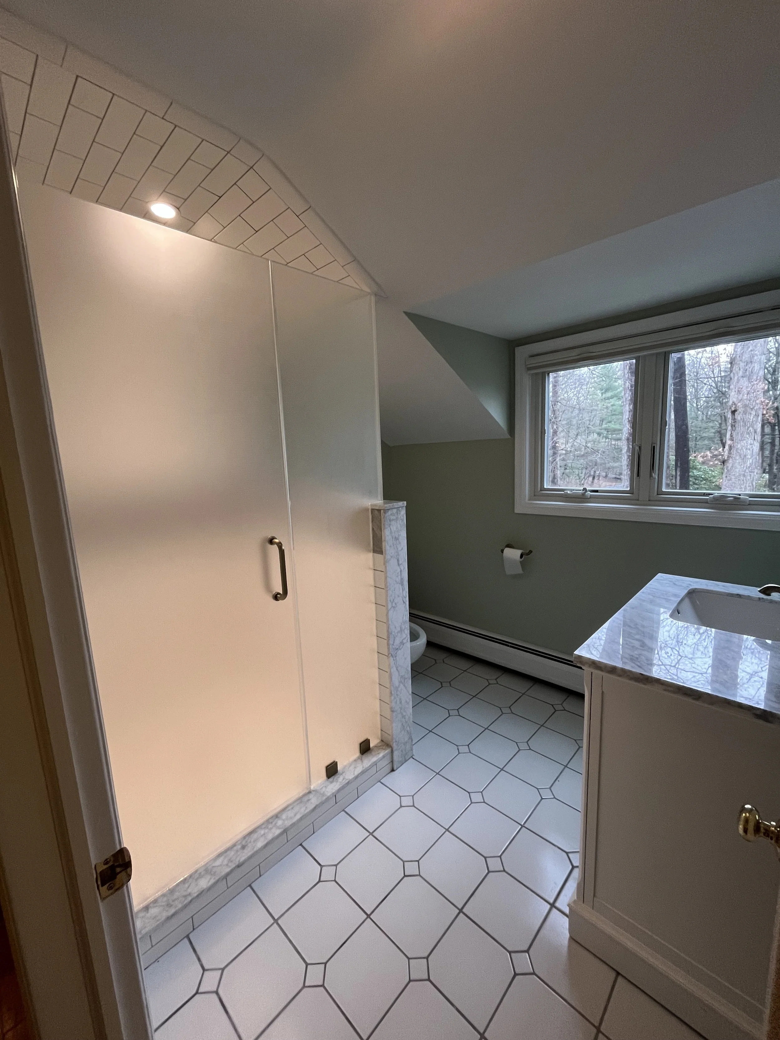 Bathroom with white tiled floor, green walls, a window showing trees outside, a sink with a marble countertop, a toilet paper holder, and a built-in shower with a handle.
