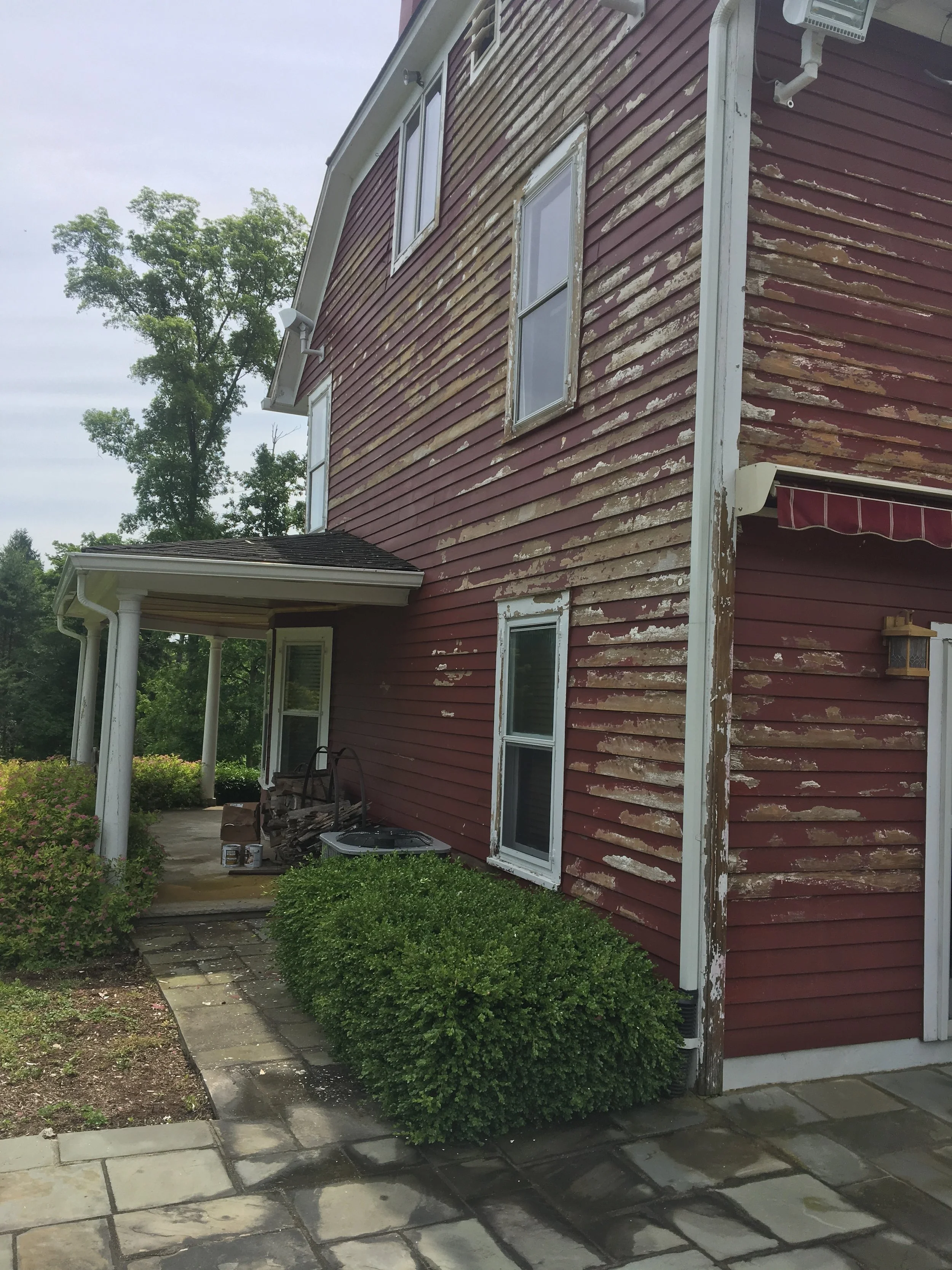 The side of a red wooden house with peeling paint, surrounded by greenery, including bushes and trees. A small porch with equipment and supplies is visible.