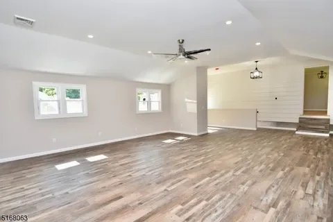 Empty living room with light hardwood flooring, white walls, three windows, ceiling fan, and a staircase leading to another level.