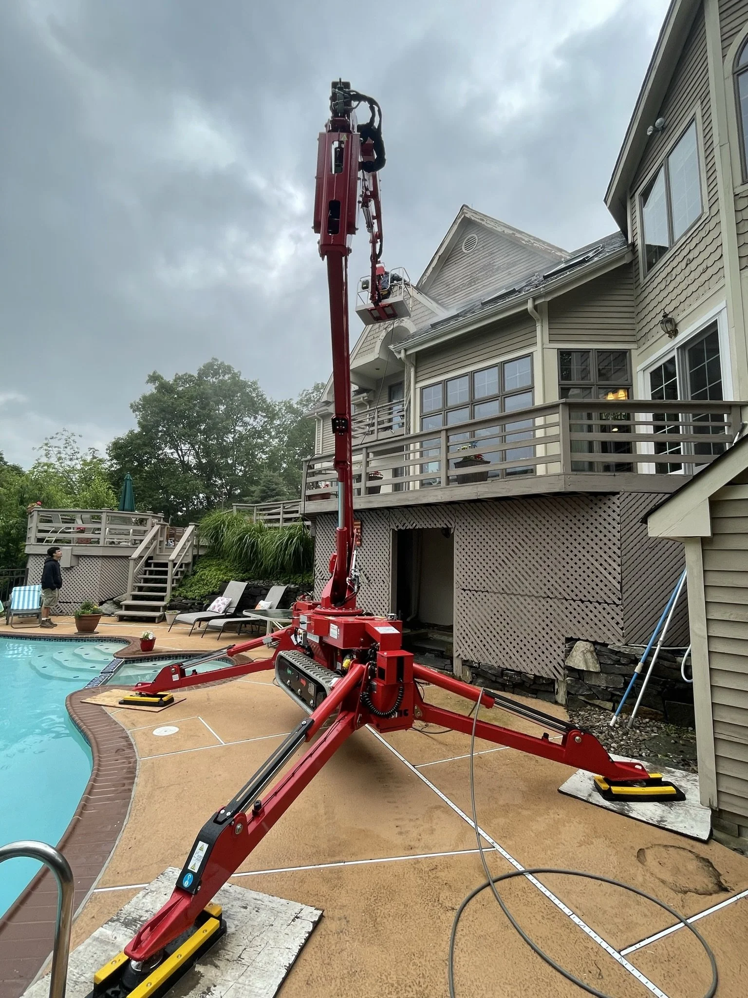 Aerial work platform lifting a worker to the roof of a house for maintenance or inspection on a cloudy day, with a swimming pool and patio furniture in the backyard.