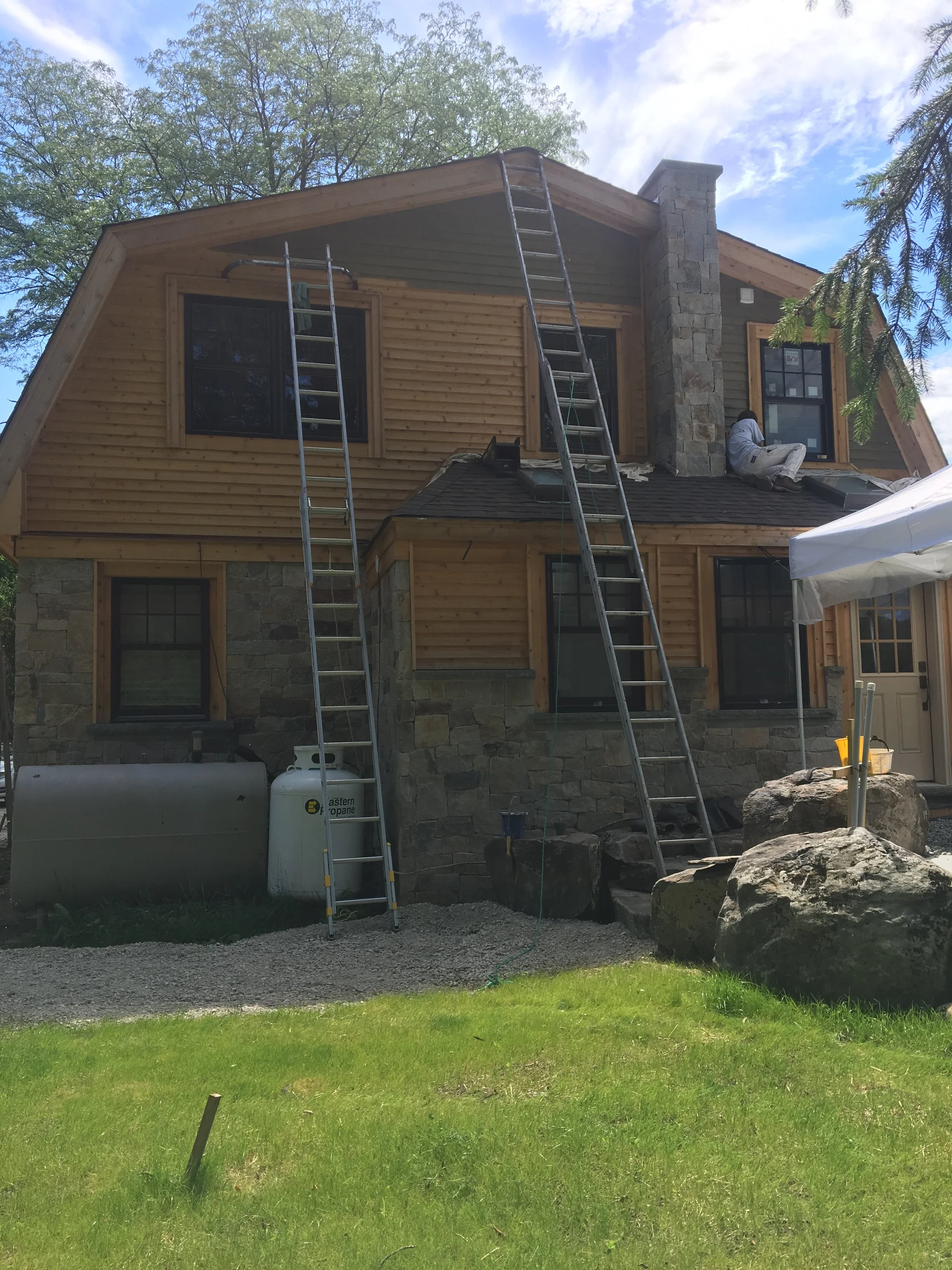 A house under construction with workers on the roof, two ladders leaning against the house, and construction materials nearby, including large rocks and a propane tank, with a grassy yard in the foreground.
