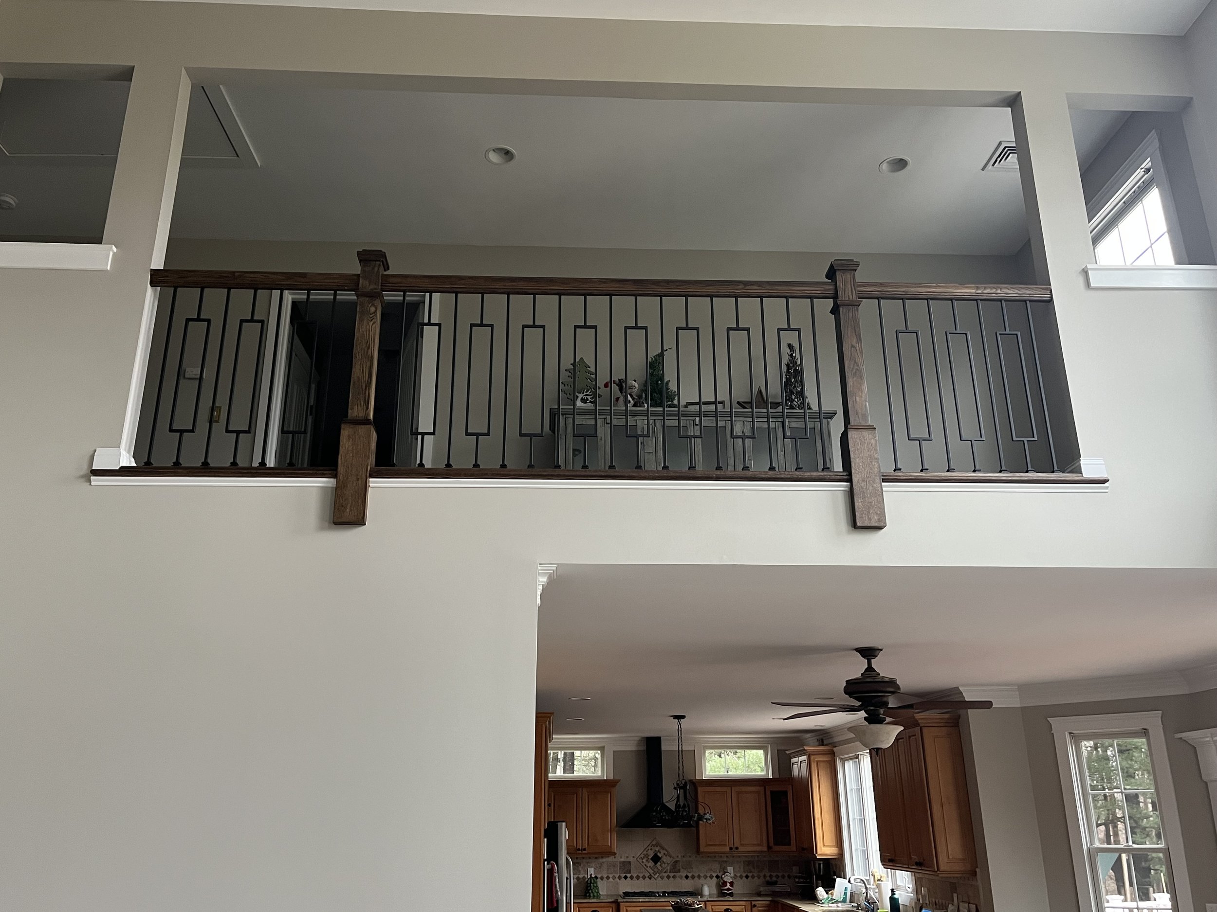 View of a two-story home interior with a second-floor balcony railing, a kitchen below with wooden cabinets, a ceiling fan, and large windows.