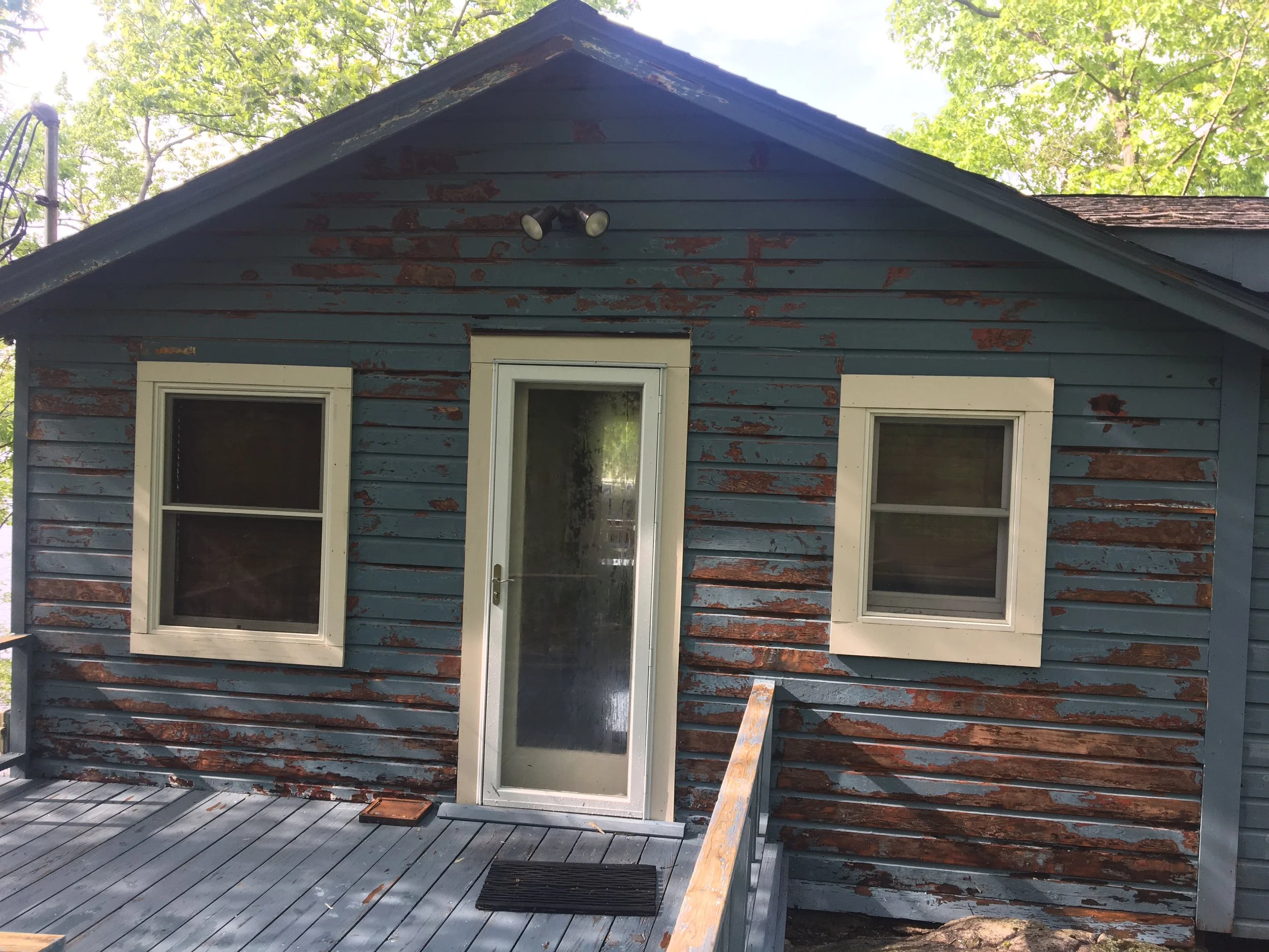 The image shows the front of an old house with blue and brown peeling paint, two windows, and a glass door, with a wooden porch and steps.