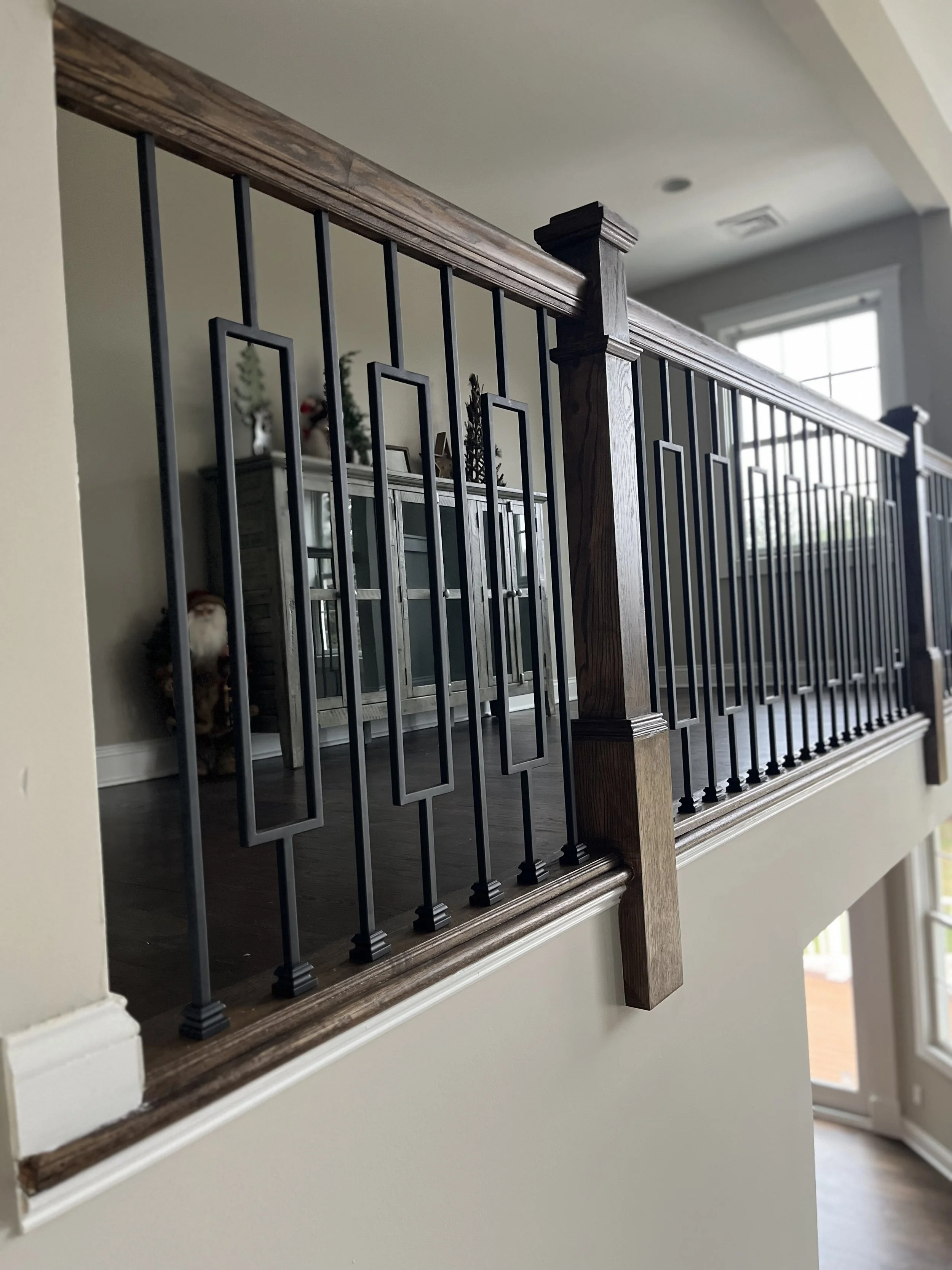 Interior view of a staircase with wooden handrail and decorative black metal balusters, overlooking a living area with Christmas decorations and a large window.