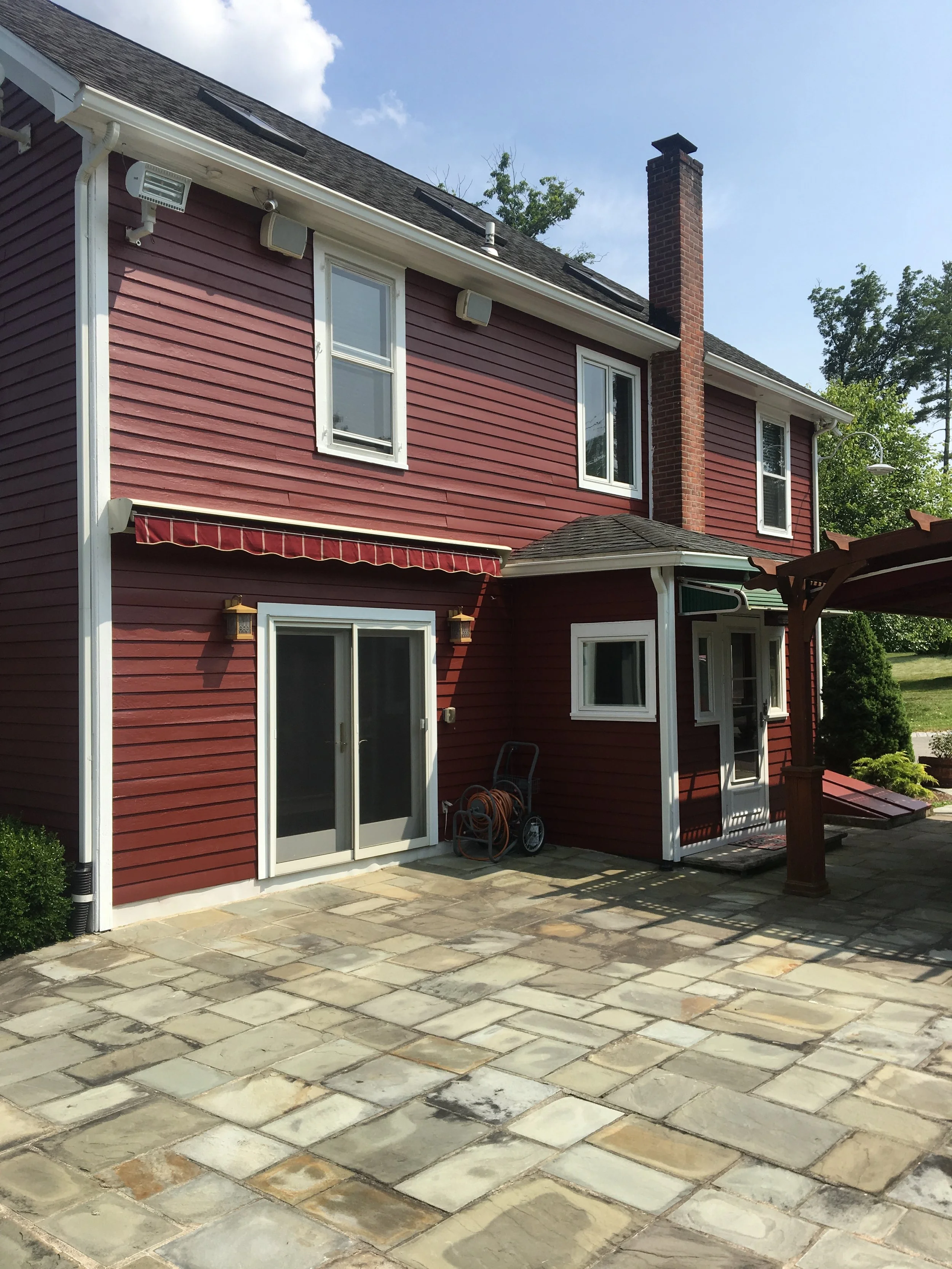 Backyard patio with stone pavers, red house with white trim, sliding glass door, small window, small porch, and outdoor hose reel.