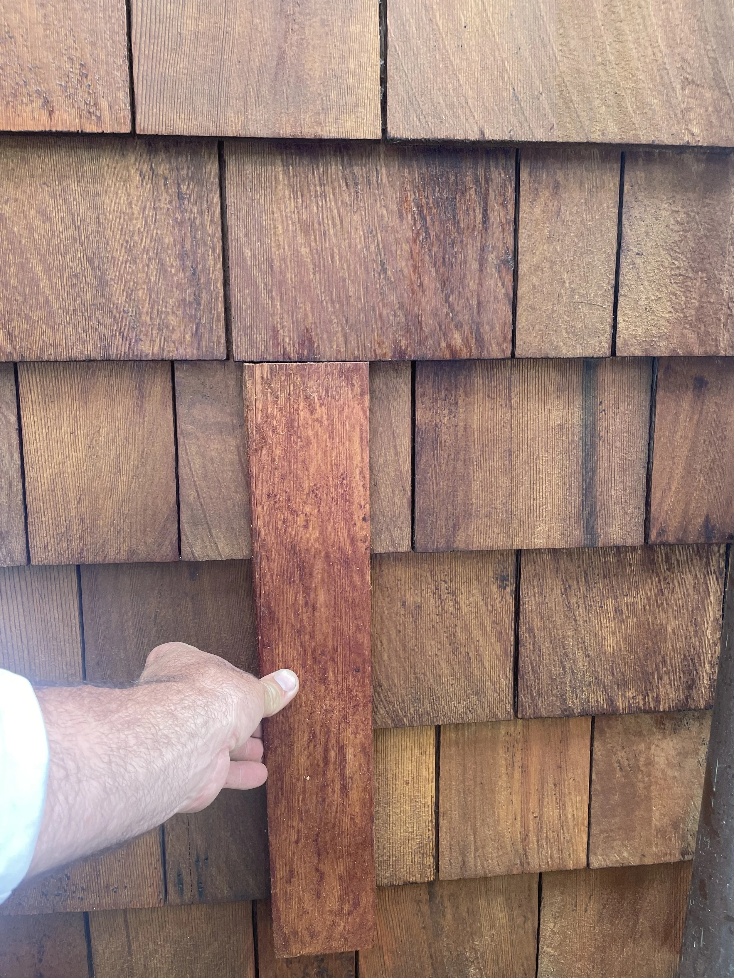 A person is holding a wooden plank in front of a wall made of cedar shingles.