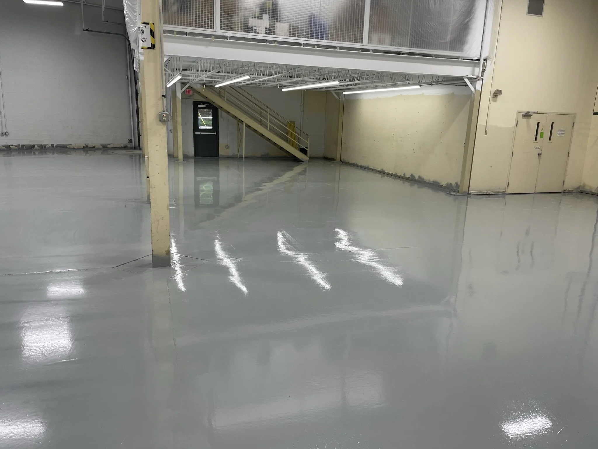 Empty indoor warehouse or industrial space with a shiny, polished concrete floor, metal support beams, stairs leading to an upper level, and a door in the background.