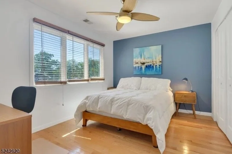 A bedroom with a wooden bed frame and white bedding, a wooden nightstand with a desk lamp, a blue accent wall with a painting, and large windows with blinds, a ceiling fan overhead, and hardwood floors.