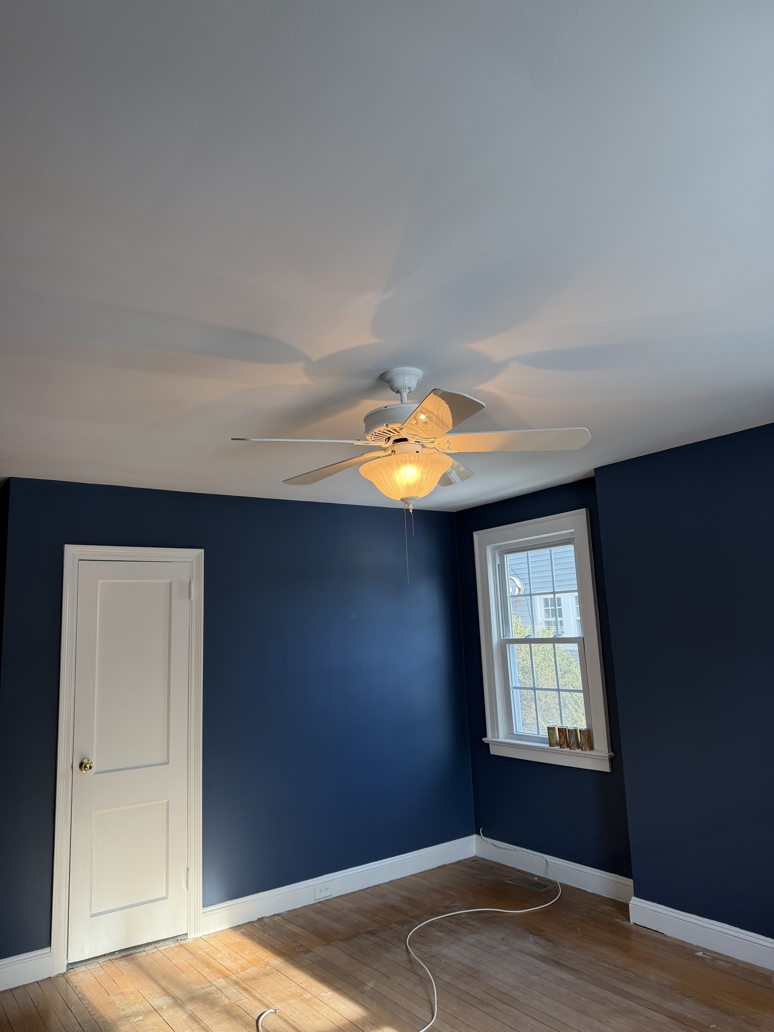 Empty room with blue walls, hardwood floors, a white ceiling fan with lights, a window showing a house outside, a white door, and a visible electrical cord on the floor.