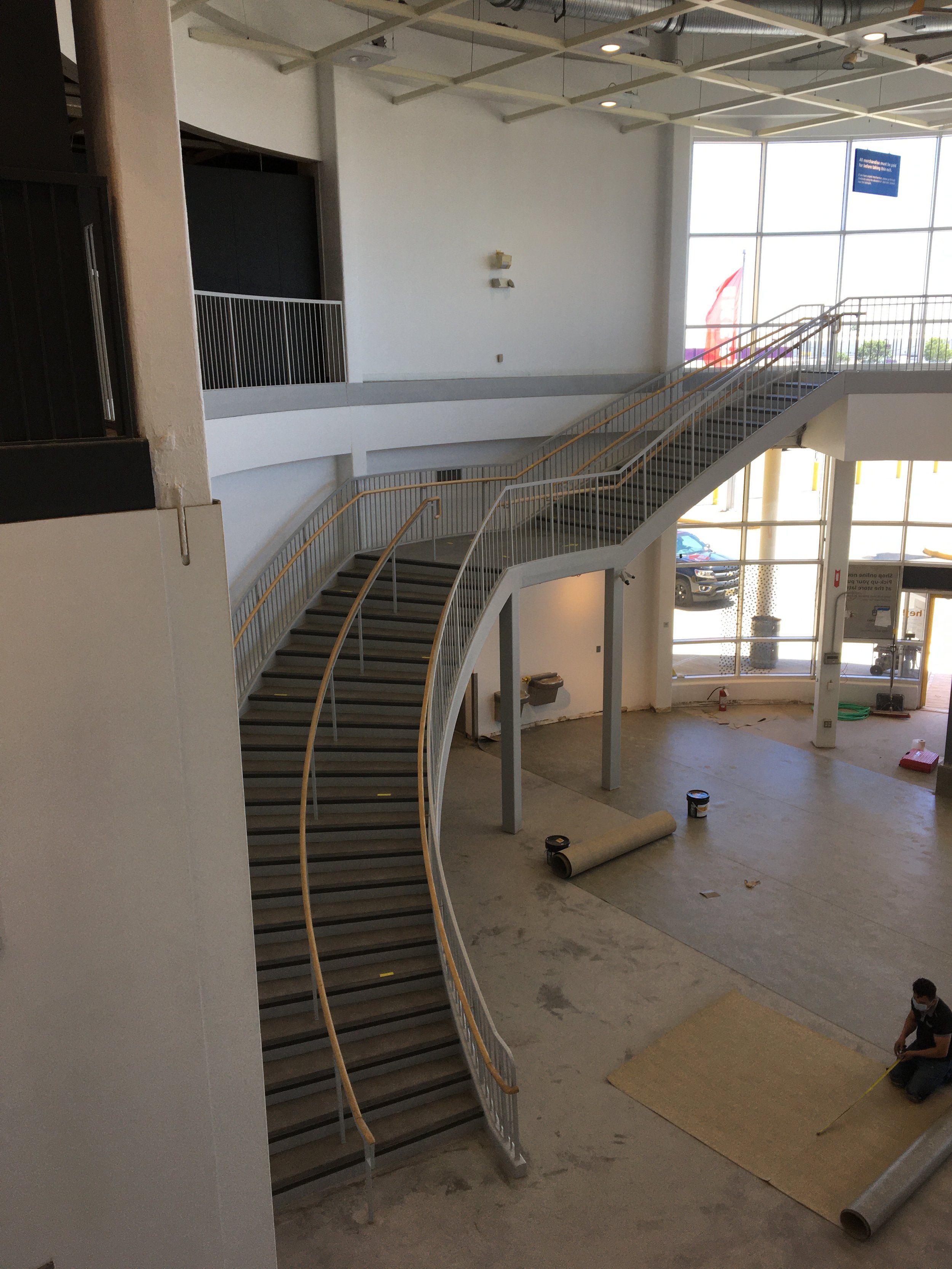 Empty commercial space with a curved staircase, large front windows, and a worker measuring on the floor during construction.