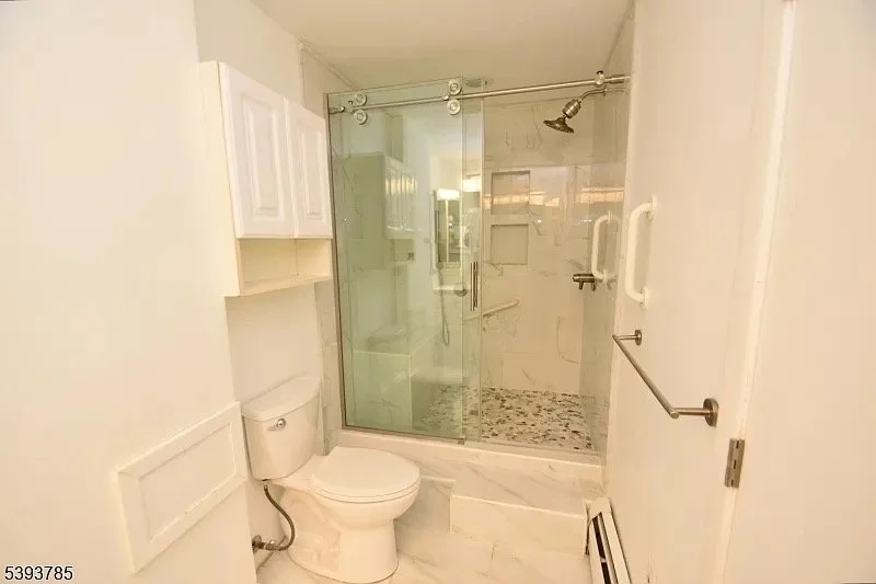 Bathroom with a toilet and a glass-enclosed shower with a sliding door, a handheld shower head, wall-mounted grab bars, and white cabinets.