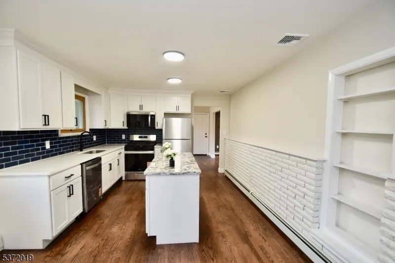 Modern kitchen with white cabinets, black tile backsplash, granite countertop island, stainless steel appliances, and hardwood flooring.